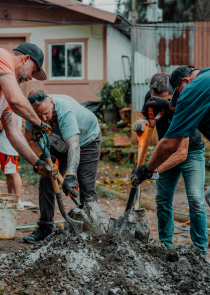 Several people outside with shovels digging in a dirt pile in front of a old house